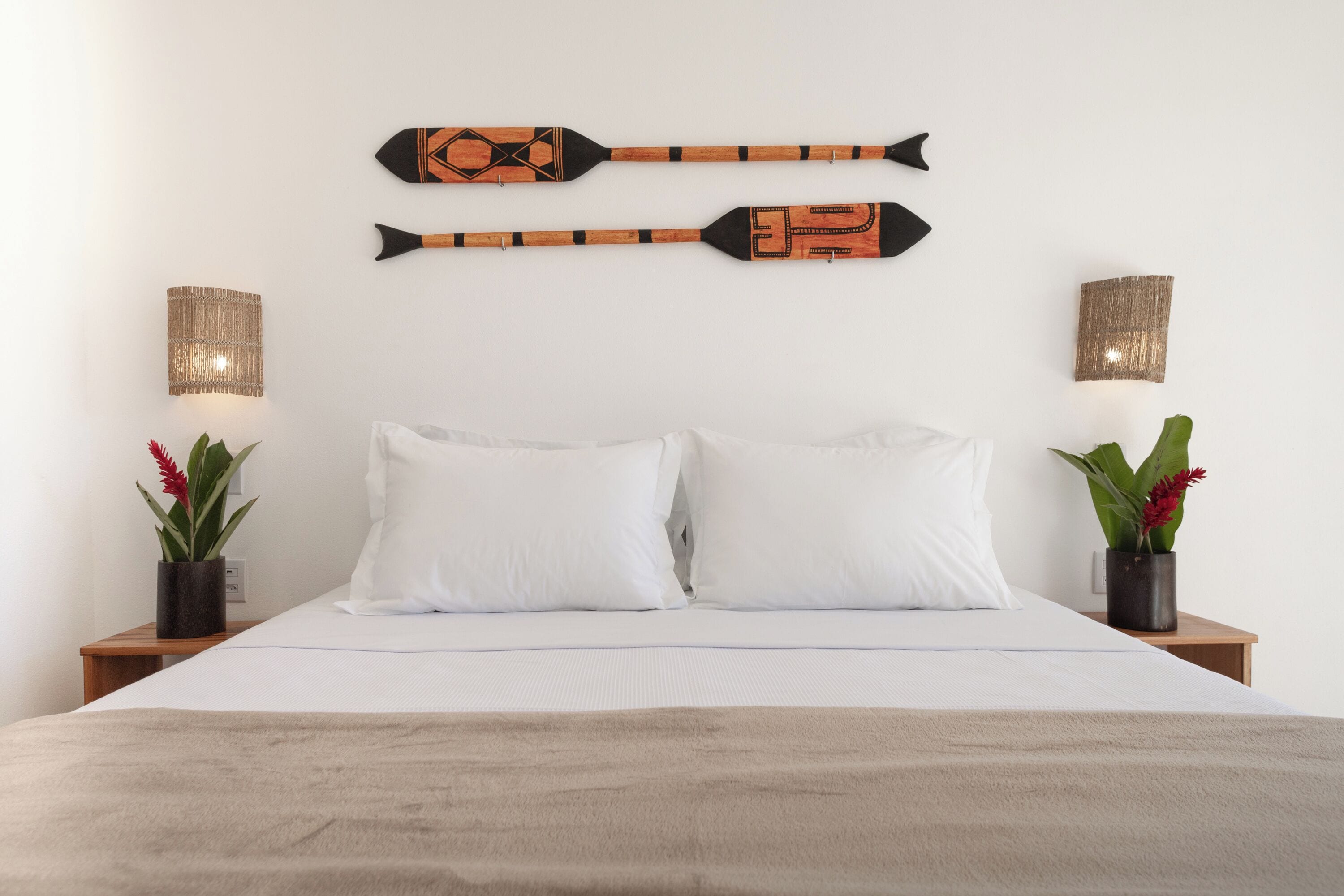 Cama com roupa de cama branca, dois remo decorativos na parede e duas mesas de cabeceira com luminárias e plantas.