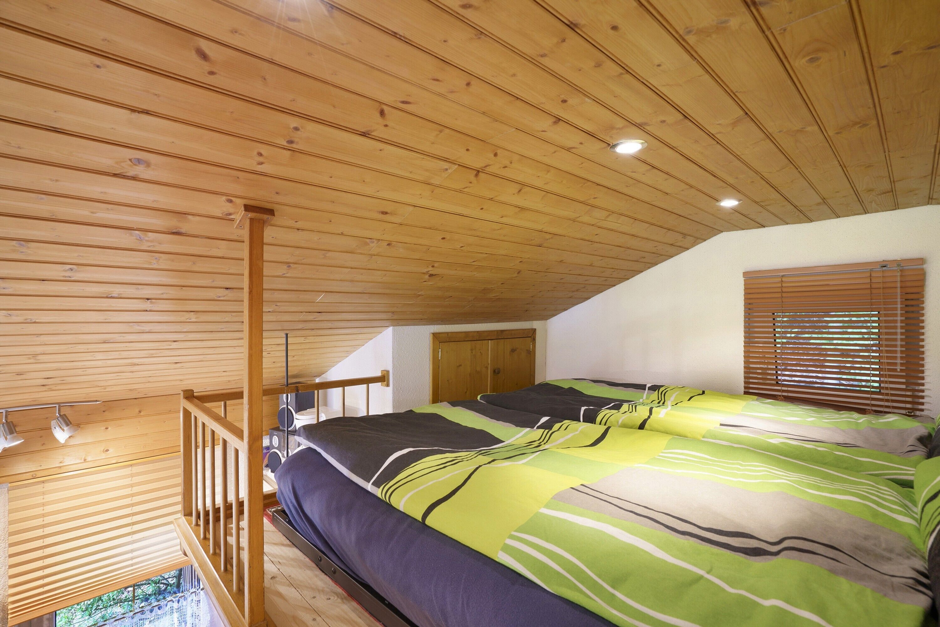 A bedroom with a sloped ceiling, a bed with striped bedding, a wooden railing, a window with blinds, and a door.