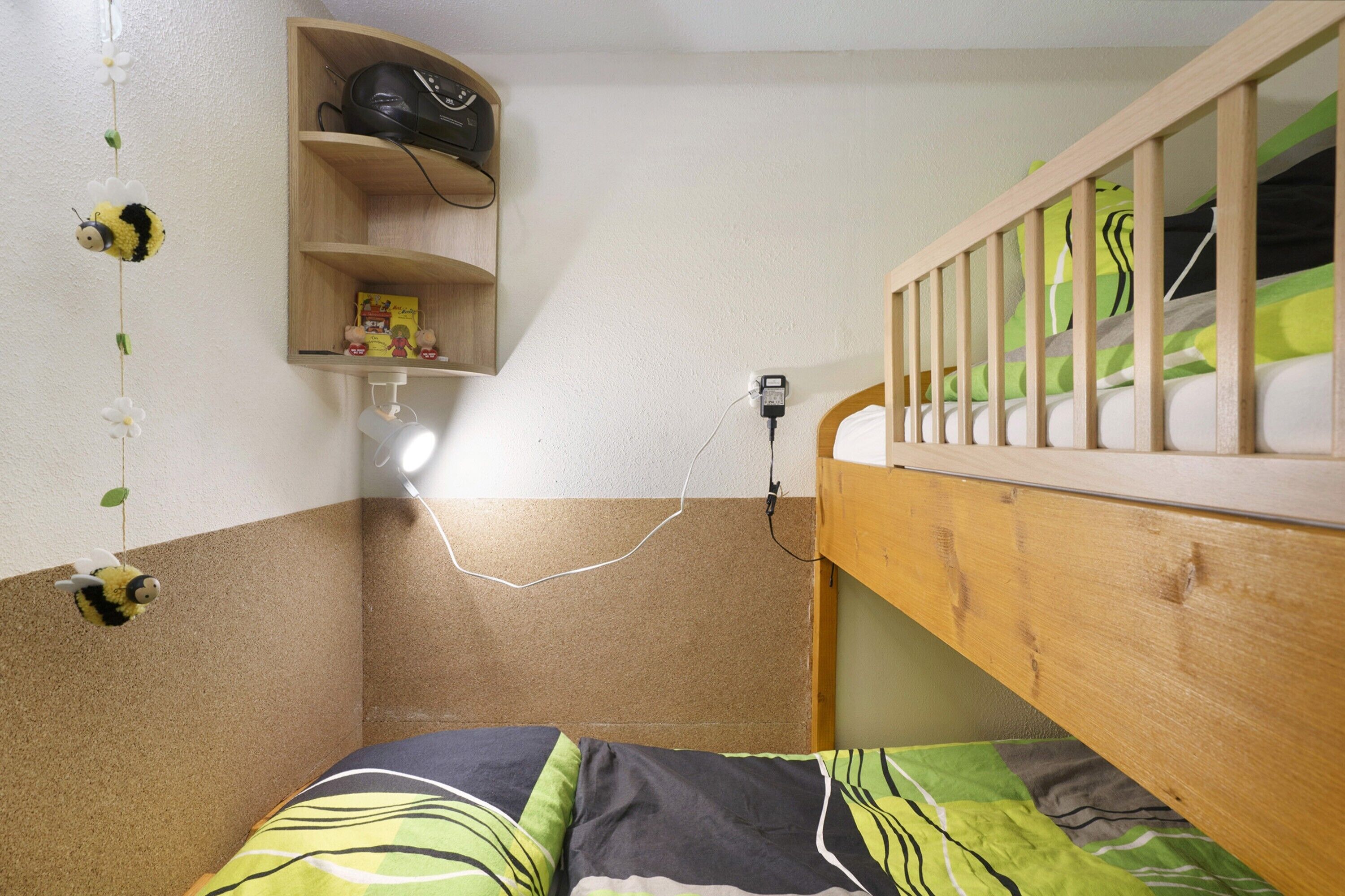 Un lit superposé avec une tête de lit en bois, une étagère sur laquelle se trouve un objet noir et une prise de courant fixée au mur.