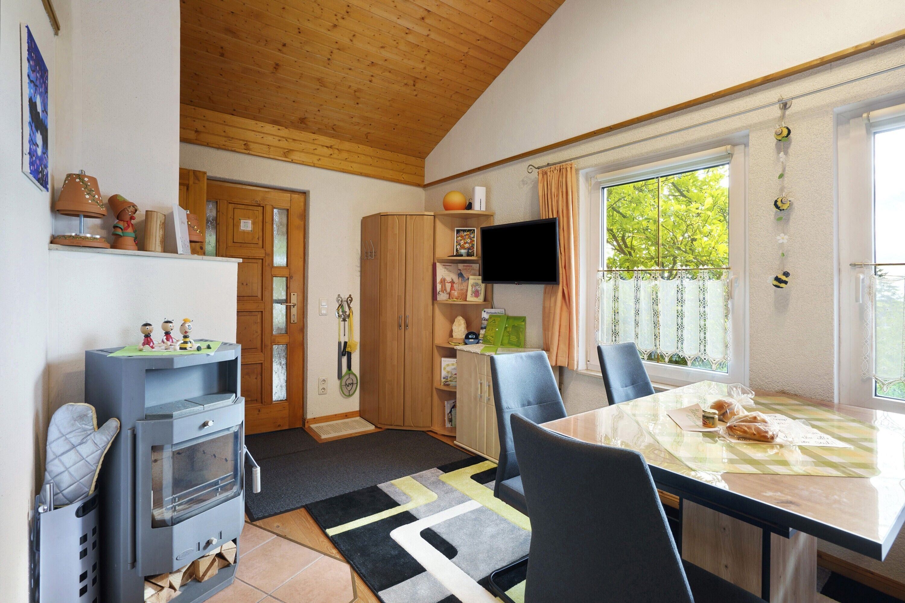 Une pièce avec un plafond en bois, une table à manger avec des chaises, un téléviseur fixé au mur et une fenêtre avec des rideaux.