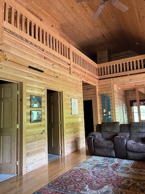A log cabin interior with a wooden ceiling, a ceiling fan, a large window, a brown leather sofa, and a patterned rug.