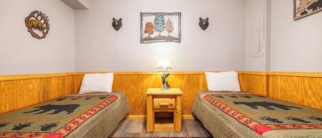 A room with two beds, a small wooden nightstand with a lamp, and decorative wall art.