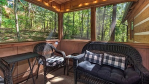 Una cabaña de madera acogedora con un porche, muebles de mimbre y ventanas amplias con vistas a un bosque.