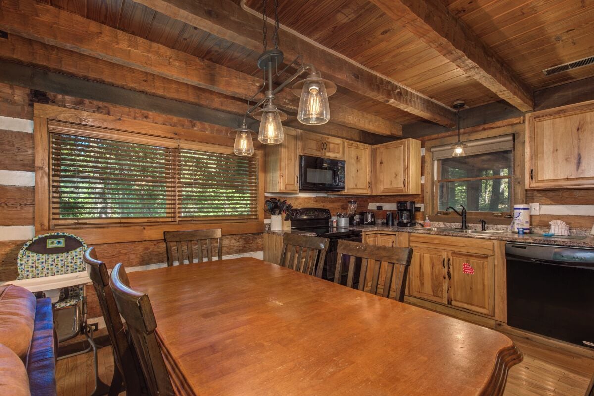 A rustic cabin interior with a wooden dining table, chairs, a kitchen with wooden cabinets, a microwave, and a large window with blinds.