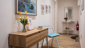 A hallway with a wooden desk, a vase of flowers, framed artwork, a blue chair, a mirror, and a white shelf.