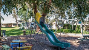A playground with a slide, picnic tables, and a tree. - Kalganyi Holiday Park (Mount Gambier)