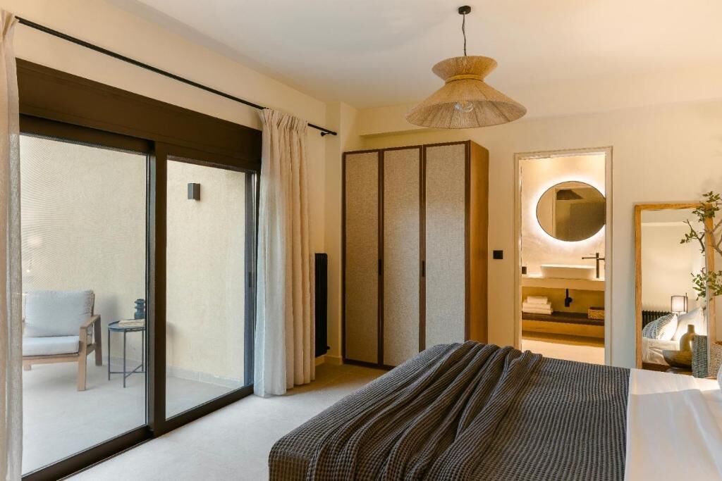 Un dormitorio moderno con una cama grande, un ventanal corredero que da a un balcón, un armario y un baño con un espejo redondo.