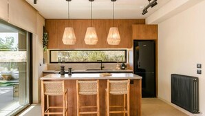 A modern kitchen with wooden cabinets, a large island with bar stools, pendant lighting, and a view through the window. - The Attican Villa & Estate (Saronikos)