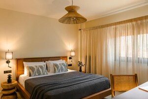 Ein Hotelzimmer mit einem hölzernen Bett, zwei Nachttischlampen, einem großen Fenster mit transparenten Vorhängen und einem Korbstuhl.