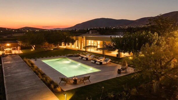 A modern outdoor pool area with lounge chairs and a covered seating area, surrounded by a landscaped garden and a view of distant hills. - The Attican Villa & Estate (Saronikos)