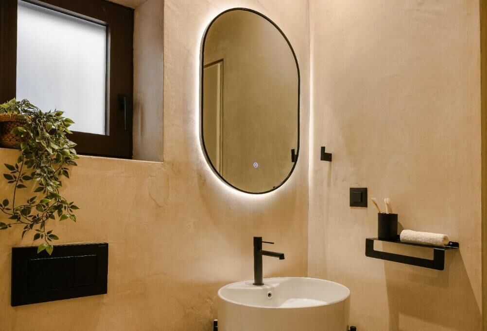 Un baño con un espejo redondo, un lavamanos blanco, una planta en maceta, un perchero para toallas y un interruptor de luz.