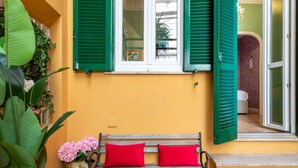 A bench with red cushions in front of a window with green shutters, a potted plant with pink flowers, and a door leading to a room with a patterned wall.