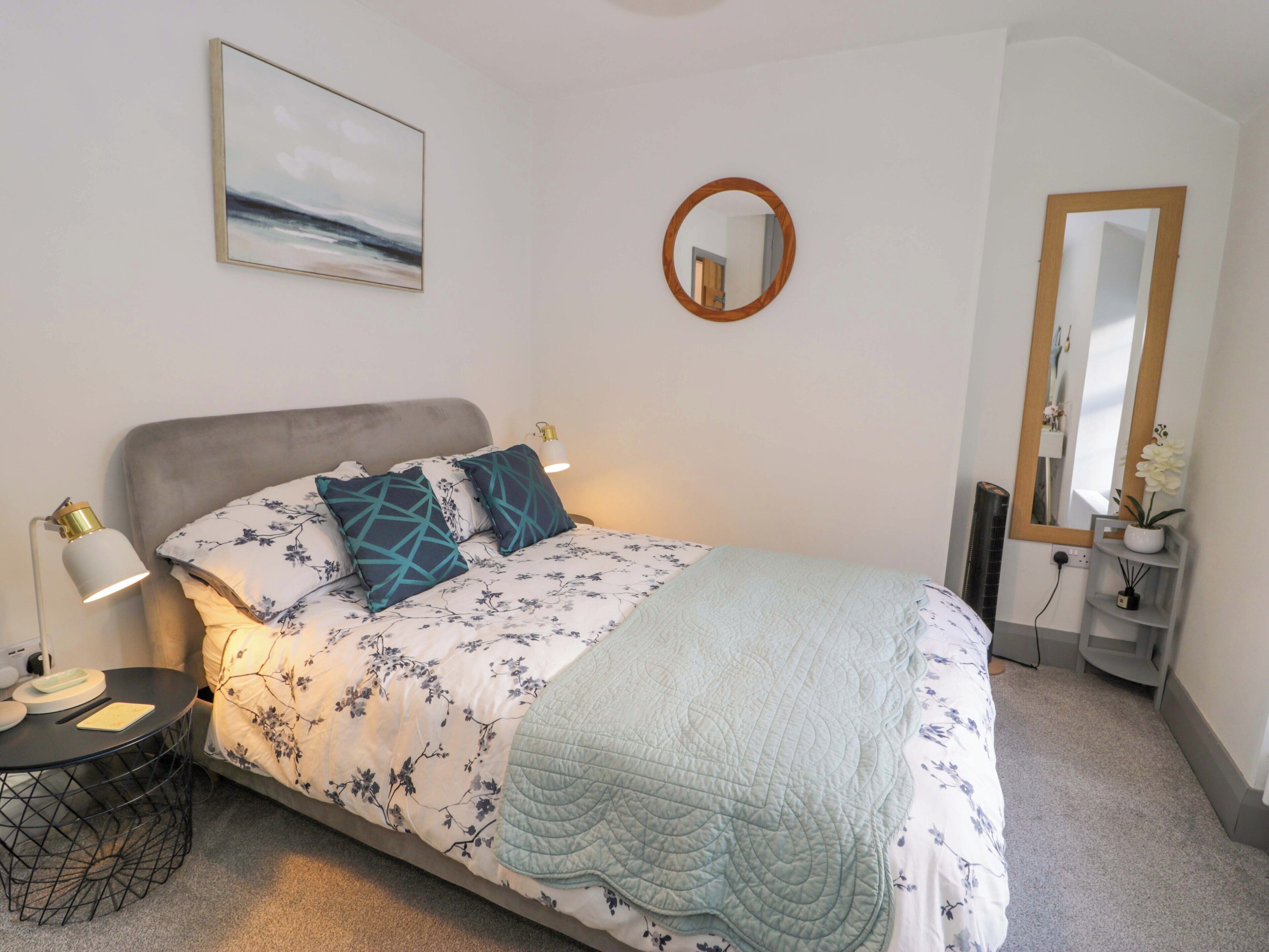 A neatly arranged bedroom with a bed, bedside tables, a round mirror, and a painting on the wall.
