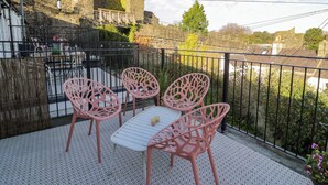 Ein Balkon mit einem weißen Tisch und rosa Stühlen, mit Blick auf ein historisches Steinhaus.