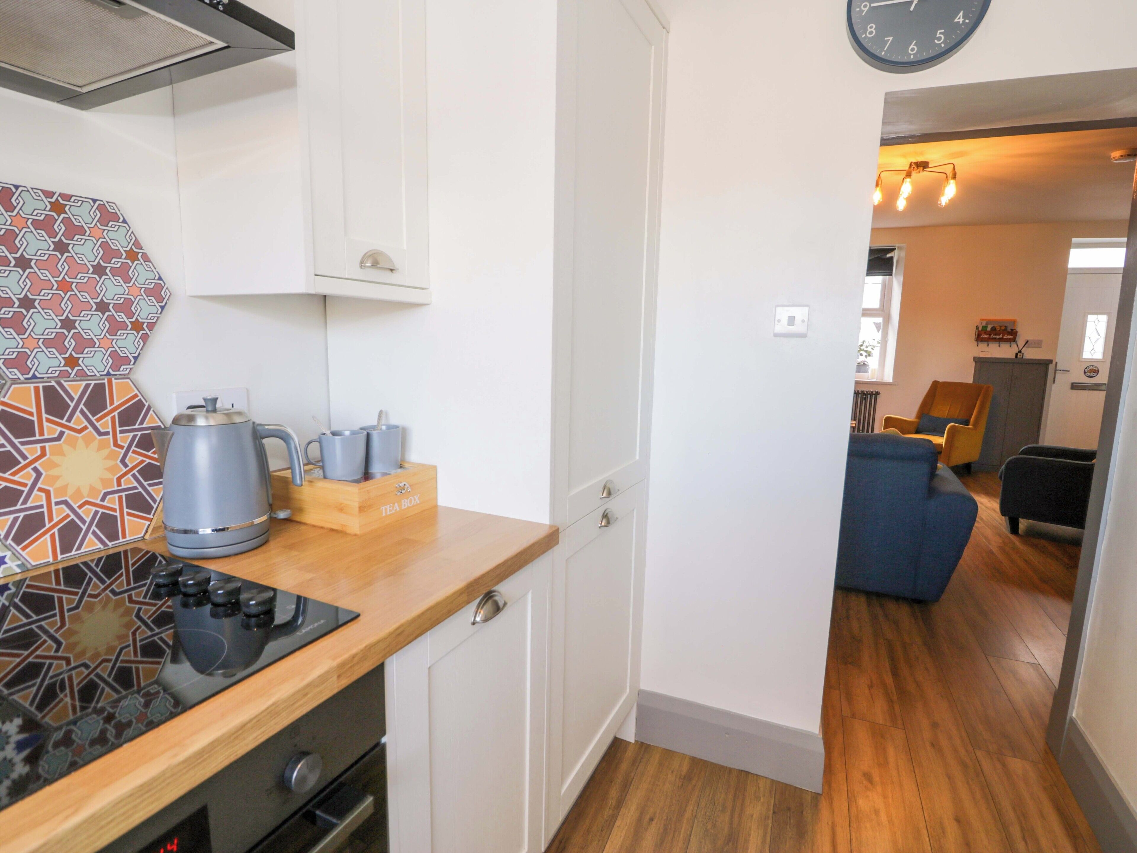 Une cuisine compacte équipée d’une cuisinière, d’une bouilloire et d’une boîte à thé, qui donne sur un espace de vie comprenant un canapé et des fauteuils.