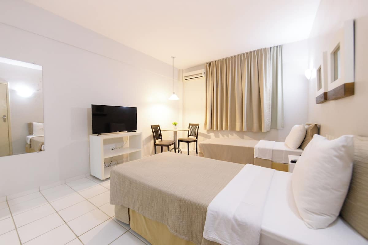 Une chambre d’hôtel comprenant un lit, une télévision, un bureau avec deux chaises, une petite table avec une plante et une fenêtre avec des rideaux.