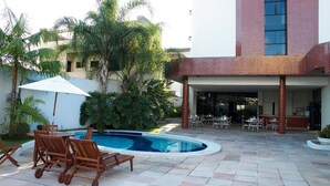 Zona de piscina exterior con sillas y mesa de madera, rodeada de palmeras y un edificio de fachada de ladrillo rojo.