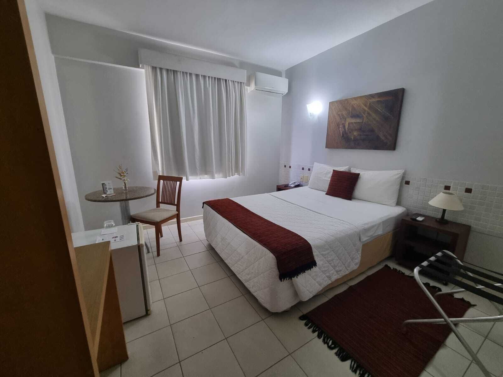 A hotel room with a bed, a desk, a chair, a window with curtains, a painting on the wall, and a lamp on a bedside table.