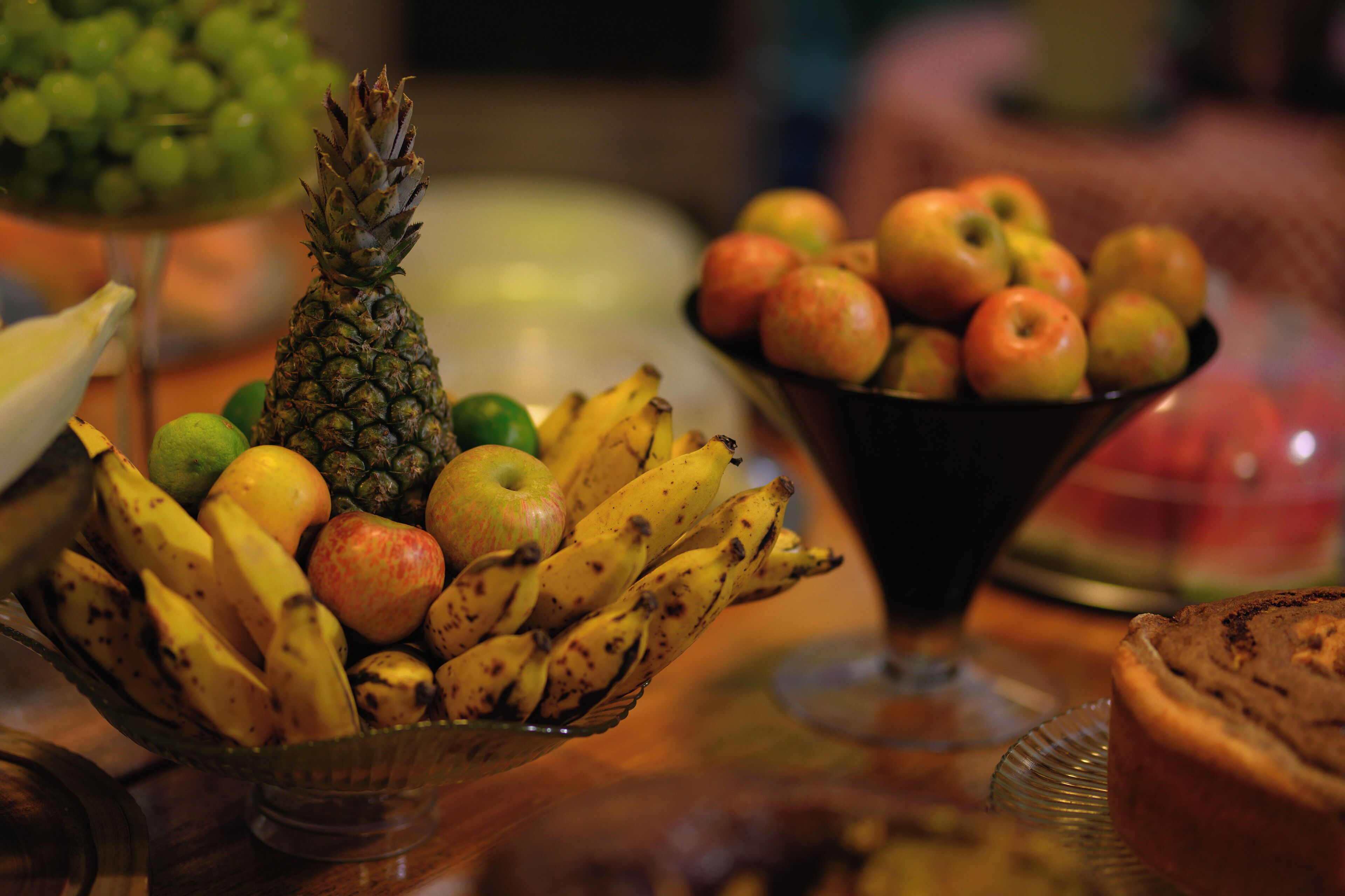 Un frutero con plátanos, manzanas y una piña sobre una mesa de madera.