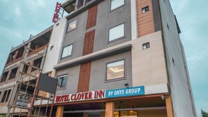 Un hotel llamado 'Hotel Clover Inn' de Onyx Group, con una fachada moderna que combina paneles de hormigón y madera, una entrada visible y varias motocicletas estacionadas afuera.