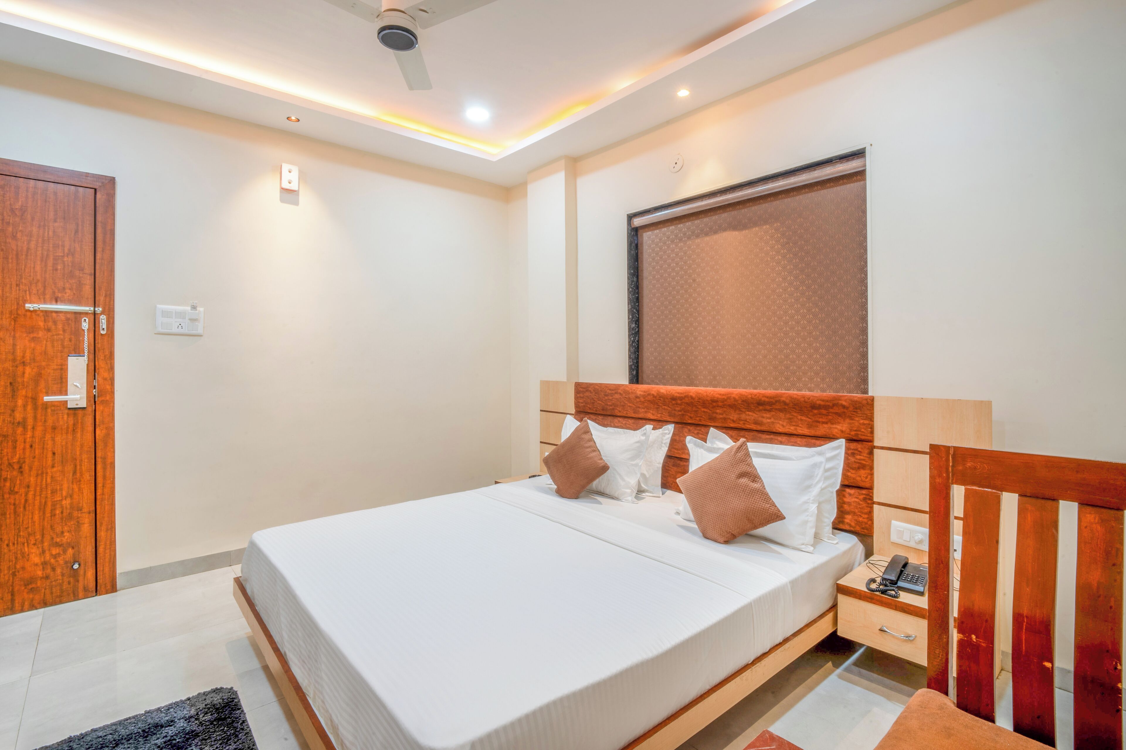 A modern bedroom with a wooden headboard, a bed with white and brown bedding, a nightstand with a phone, and a ceiling fan.