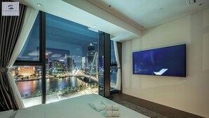 Habitación de hotel con una cama grande, un televisor de pantalla plana y vistas a la ciudad a través de amplios ventanales.