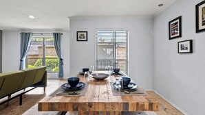 A dining area with a wooden table set for four, a green sofa, and a window with blinds. - Spacious Preston Hollow Condo (Dallas)