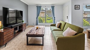 A living room with a flat-screen TV, a wooden coffee table, a green sofa, and a view of greenery outside. - Spacious Preston Hollow Condo (Dallas)