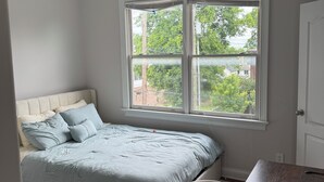 A bedroom with a bed, a desk, a chair, a ceiling fan, and a window with a view of trees and houses. - Bright & Airy 2-bed, 2.5-bath house in historic Germantown, Nashville (Nashville)