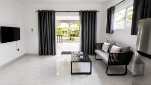 A modern living room with a flat-screen TV, a sofa with cushions, a coffee table, and a large window with curtains. - Trwa Koko Self Catering (Praslin Island)