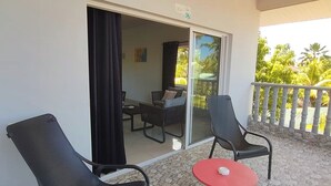 Ein Balkon mit zwei Stühlen und einem kleinen Tisch, eine Schiebetür zu einem Innenbereich mit einer Couch und einem Tisch, und Blick ins Grüne.