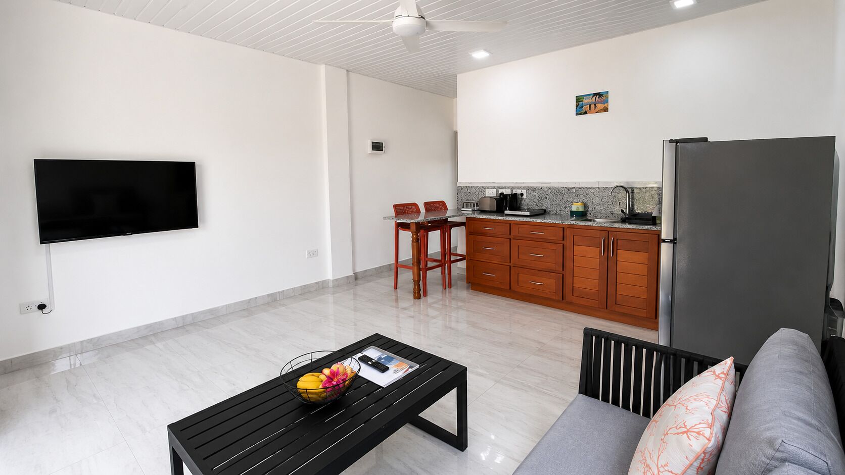 A modern living space with a flat-screen TV, a dining area with chairs, a kitchen with wooden cabinets, and a grey sofa.