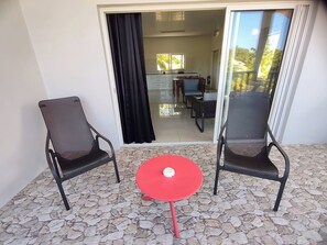 Ein Balkon mit zwei schwarzen Korbmöbeln, einem roten Tisch und einer Glastür, die zu einem Innenbereich mit Esstisch und Stühlen führt.