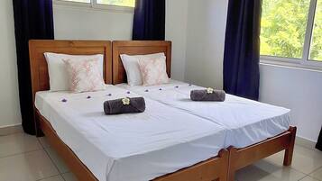 Un lit double avec un cadre en bois, une literie blanche et des coussins décoratifs. Un climatiseur est fixé au mur, et la pièce est dotée de grandes fenêtres avec des rideaux.