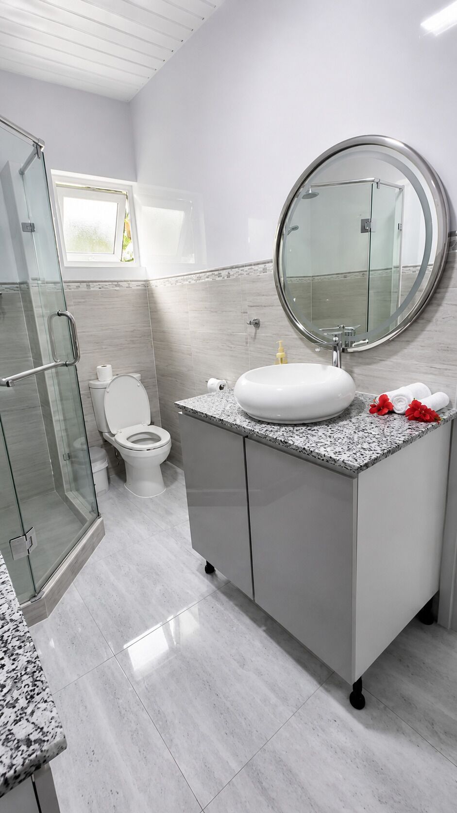 Un baño moderno con cabina de ducha de vidrio, inodoro blanco, espejo redondo y encimera de granito.