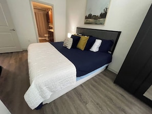 A bedroom with a bed, a nightstand, a painting on the wall, and a door leading to a bathroom.