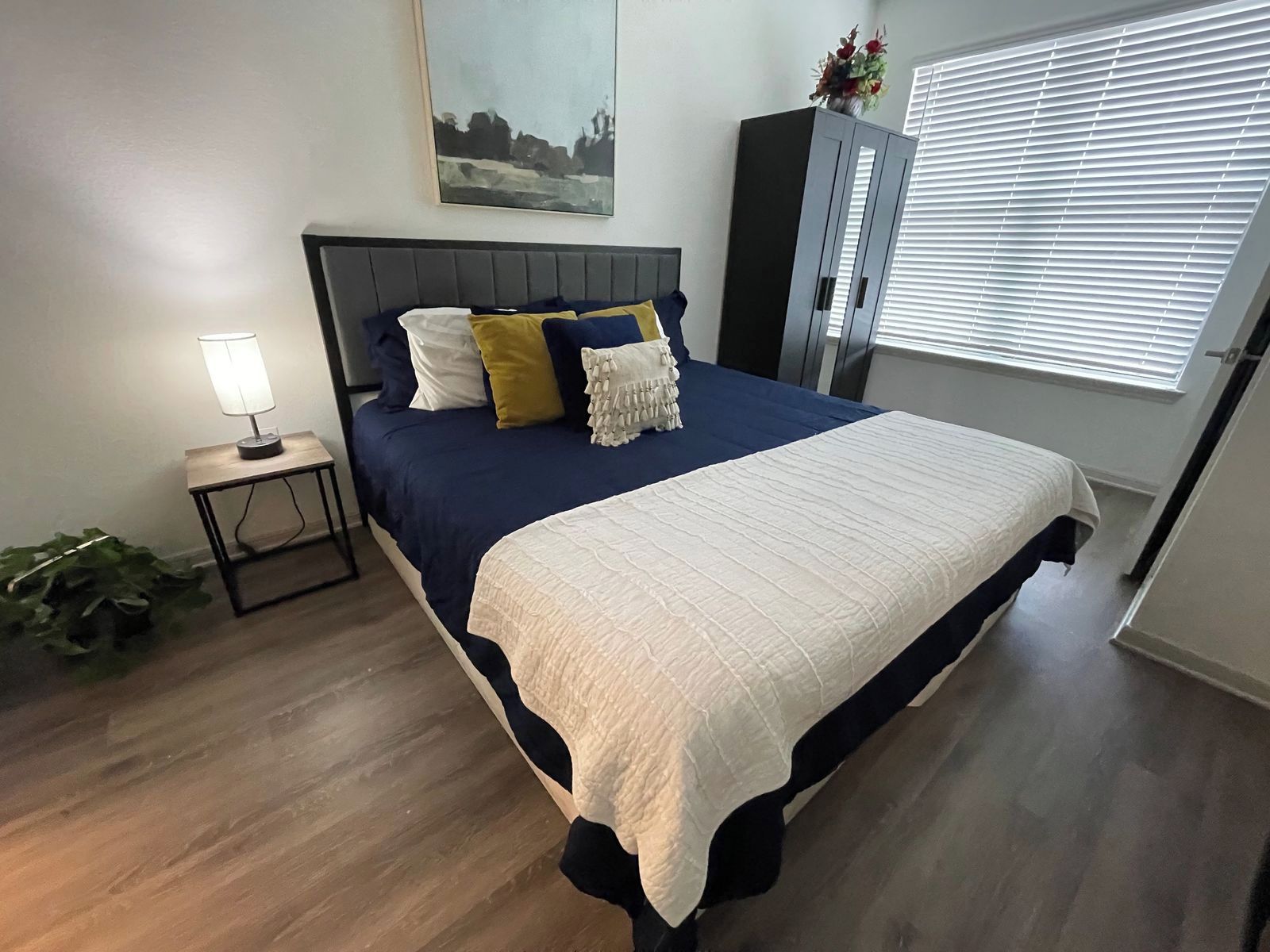 A bedroom with a bed, a nightstand, a lamp, a window with blinds, and a cabinet.