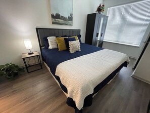 Un dormitorio con cama, mesita de noche, lámpara, ventana con persianas y armario.