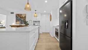 Una cocina moderna con armarios blancos, una isla grande, electrodomésticos de acero inoxidable, iluminación colgante y un cuadro abstracto y colorido en la pared.