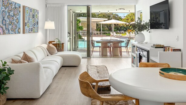 Una sala de estar moderna con un sofá, una mesa de comedor y un televisor. Se ve un área de piscina exterior a través de las puertas de vidrio.