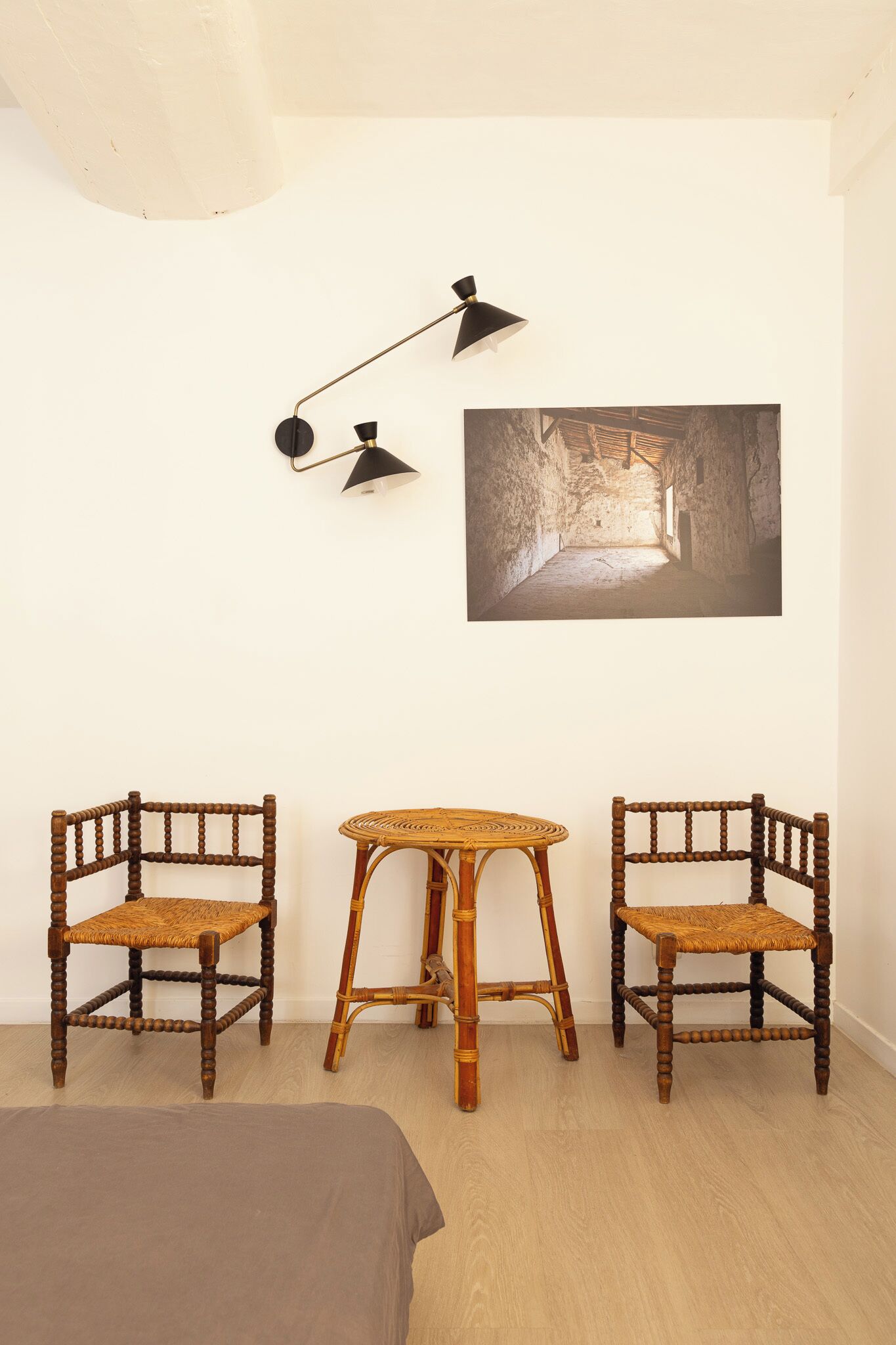 Ein Zimmer mit zwei Holzstühlen, einem kleinen runden Tisch, einem Bett und einer Wandlampe.