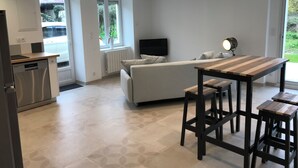 Eine moderne Küche mit Geschirrspüler, ein Essbereich mit Holztisch und Hockern, eine Couch und ein an der Wand befestigter Fernseher.