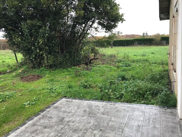 Une terrasse en bois mène à une zone herbeuse, avec un grand arbre et une haie en arrière-plan.