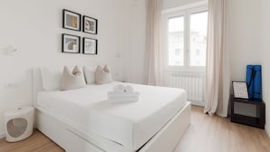 A modern bedroom with a large bed, a window with curtains, a small bedside table, and a wall-mounted picture frame.