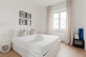 A modern bedroom with a large bed, a window with curtains, a small bedside table, and a wall-mounted picture frame.