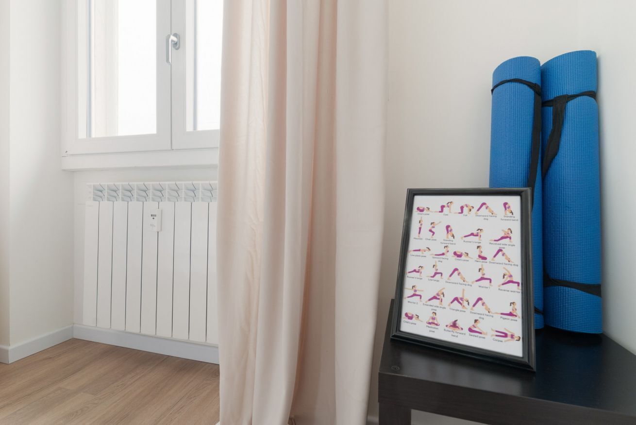 Ein Zimmer mit einer gerahmten Yoga-Matte-Übungsübersicht, einem Fenster mit Vorhängen und auf einem Tisch gerollten blauen Yogamatten.