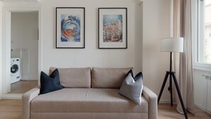 A living room with a beige sofa, two framed artworks, a floor lamp, and a laundry area.