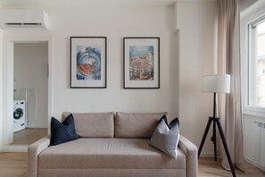 A living room with a beige sofa, two framed artworks, a floor lamp, and a laundry area.