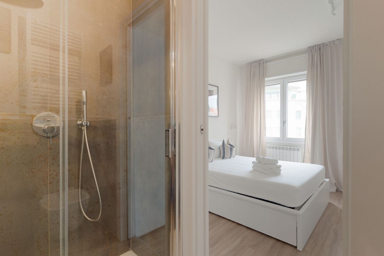 Una habitación de hotel moderna con ducha de vidrio, una cama bien hecha y una ventana con cortinas.