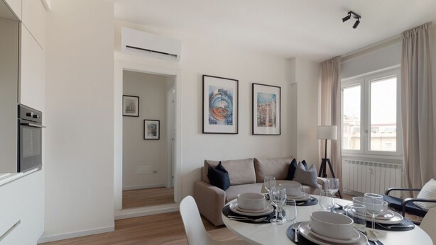 A modern dining area with a round table set for four, a sofa, and a kitchen area with an oven.